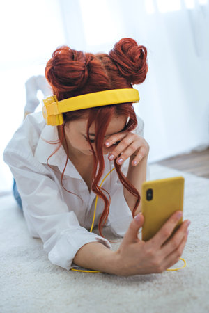 A girl sits on the bed, looks out the window and listens to music in yellow headphones.の写真素材