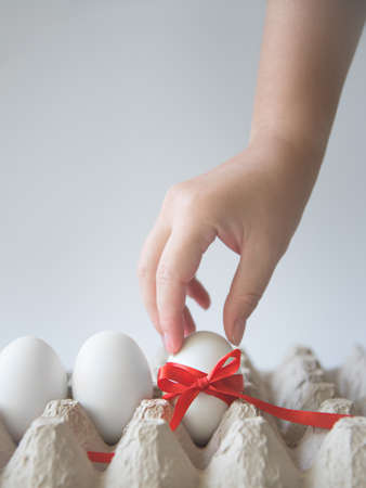 Womans hand takes white egg with red gift ribbon in an egg carton on a white background.の写真素材