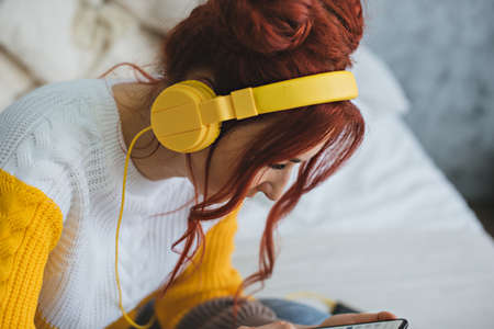 A girl with red bright hair lies on the bed and listens to music in yellow headphones.の写真素材