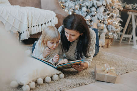 Mom and daughter are reading a book lying on the carpet near the Christmas treeの写真素材