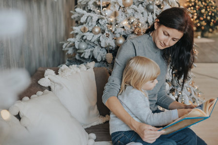 Mom and daughter are reading a book near the Christmas tree