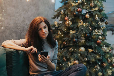 Woman chatting on social networks, messengers on the phone, sitting on the couch near the Christmas tree.の写真素材