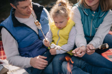 A family in brightly colored waistcoats fries marshmallows on sticks by the fire. Blurred, selective focus on marshmallows.の写真素材