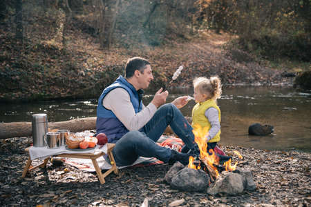 Dad and daughter in brightly colored waistcoats fries marshmallows on sticks by the fire.の写真素材