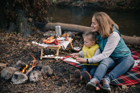 Mom and daughter in brightly colored waistcoats fries marshmallows on sticks by the fire.の写真素材