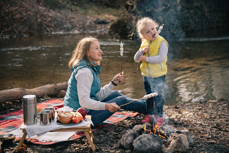 Mom and daughter in brightly colored waistcoats fries marshmallows on sticks by the fire.の写真素材