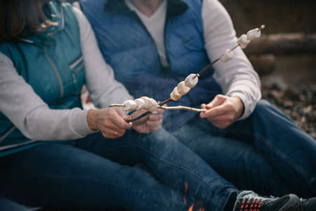 Couple in brightly colored waistcoats fries marshmallows on sticks by the fire. Blurred, selective focus on marshmallows.の写真素材