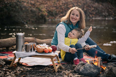 Mom and daughter in brightly colored waistcoats fries marshmallows on sticks by the fire.の写真素材