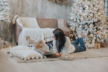Mom and daughter are reading a book while lying on the carpet near the Christmas treeの写真素材