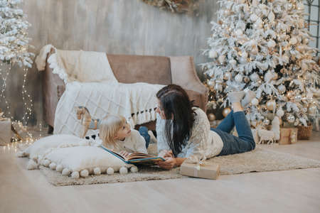 Mom and daughter are reading a book while lying on the carpet near the Christmas treeの素材 [FY310178712107]