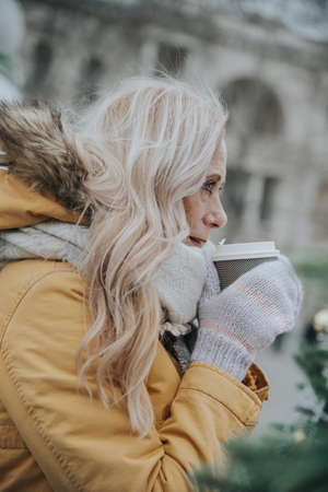 Aged pretty woman with blond hair drinks coffee in the square among the Christmas trees.の写真素材