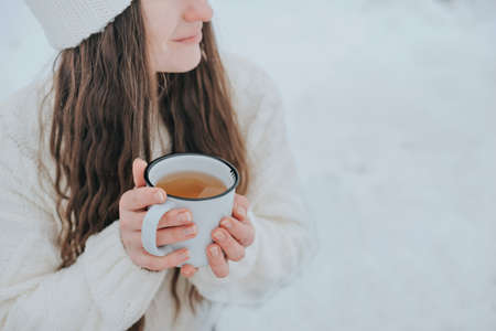 A woman in a white knitted sweater holds a white mug with a hot drink in her hands. outside in winter.の写真素材