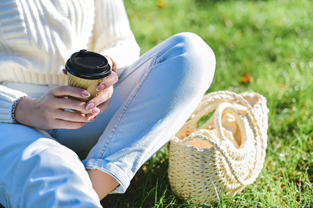 A woman in a white sweater and blue jeans sits on the grass and holds a glass of coffee in her hands.の写真素材