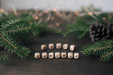 Wooden cubes with letters make up the phrase Merry Christmas on a wooden table. Fir branches, cones and a festive garland in the background.の写真素材