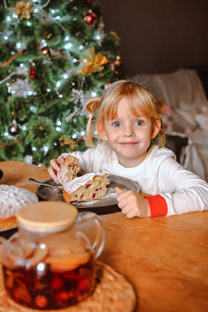 A girl with blond hair in pajamas eats a traditional Christmas cake at a table near the New Year treeの写真素材