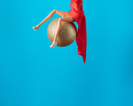 Girl doll in red dress swinging on gold Christmas bauble decoration on blue background. Wrecking ball concept. New Year inspiration.の写真素材
