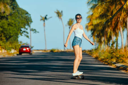 Outdoor summer portrait of cheerful young woman riding longboard in sunny weatherの写真素材