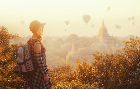 woman traveler with backpack enjoying views of Buddhist stupas and air balloons in ancient Bagan...