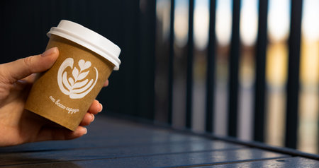 A womans hand holds a paper cup with coffee with a white lid