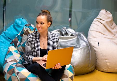 A young girl is sitting on a soft pouf with a laptop, working.の写真素材