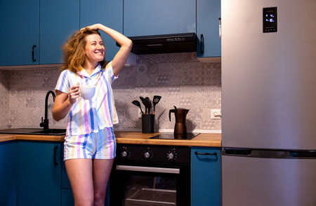 young woman pajamas holding mug and fixing her hair in the kitchen. Smiling