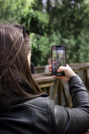 young girl holds phone her hands takes pictures of nature. View from the girls shoulder. Dark hair,...
