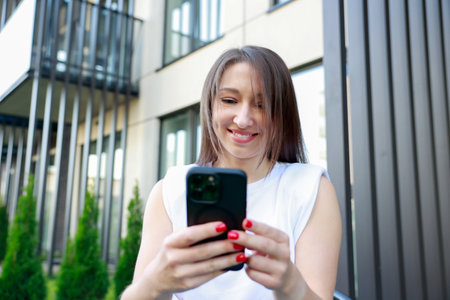 Young woman sitting outdoors modern urban setting, focused smartphone Remote work conceptの写真素材