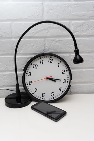 A black desk lamp next to a clock and a mobile phone. Black mobile phone. Watch with red second hand