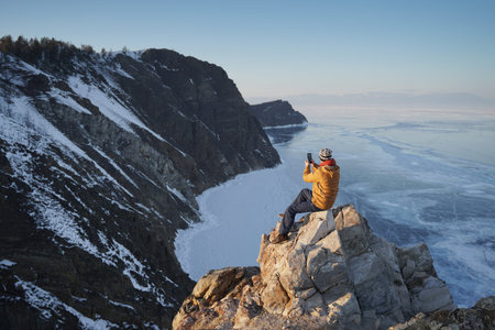 Lake Baikal at winter. Man sitting on a cliff and taking photo on smartphone. Frozen Baikal lake....