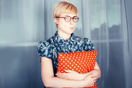 Lifestyle portrait of a beautiful blonde girl in glasses sitting on window sill and holding red...