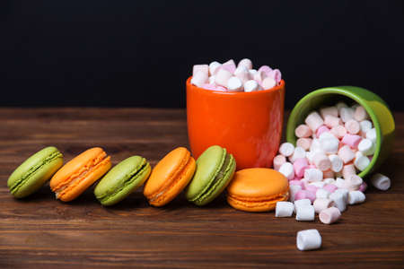 Two cups orange and green full of marshmallow with french pistachios and orange macaroons in front in dark colors on a wooden background. Shallow depth of fieldの写真素材