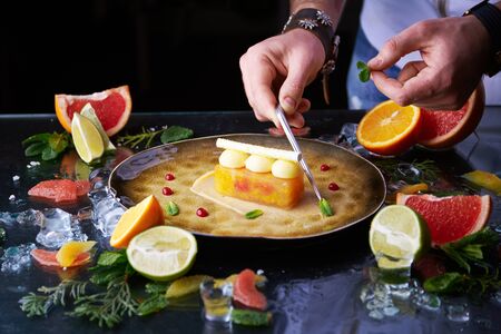 Chef hands putting mint leaves around citrus tart on a plate with a lot of citrus fruits and ice aroundの写真素材