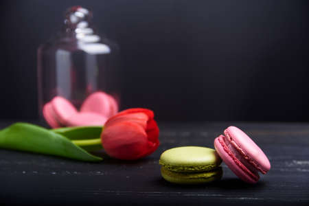 Tasty sweet pink and green macaroons on a black wooden table with red tulip and bunch of macaroons under the glass at the backの写真素材