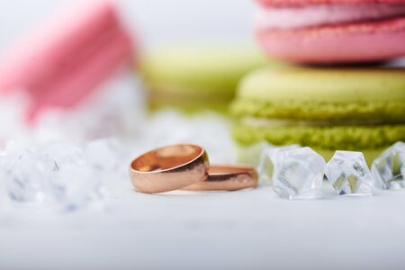 Two wedding rings lying around scattered diamonds and sweet macaroons on a white wooden backgroundの写真素材