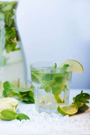 Glasses of a cold fresh lemonade drink with slices of lime on the glass, and decanter Slices of lime, ice and mint leaves on a  white wooden background. Copy space. Vertical shotの写真素材