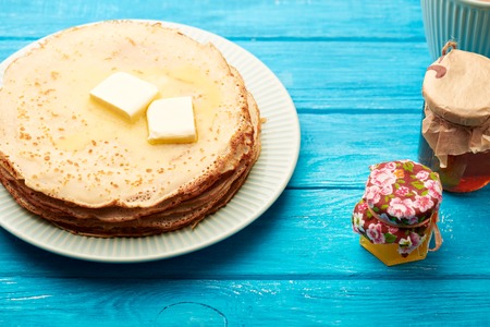 Pancakes with butter and jam jars on the blue table in front of a window and cups at the backgroundの写真素材