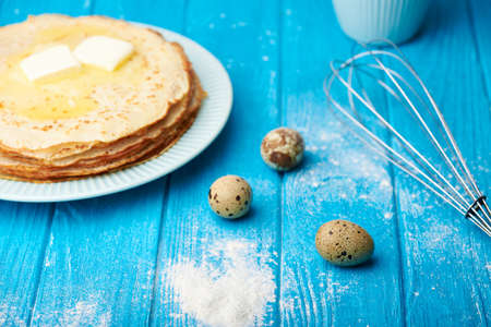Preparing to cook pancakes. Flour, whisk for whipping and quail eggs all over blue wooden table. Cooked stacked of tasty pancakes on a background. Selective focus.の写真素材