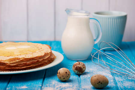 Preparing to cook pancakes. Flour, whisk for whipping, decanter with milk  and quail eggs all over blue wooden table. Cooked stacked of tasty pancakes on a background. Selective focus.の写真素材