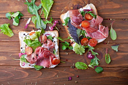 Top view at sandwich with prosciutto, tomato, salad and soft cheese on a wooden rustic backgroundの写真素材
