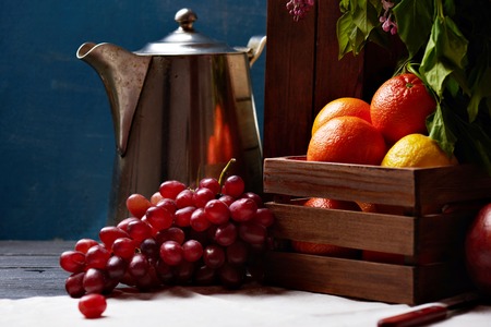 Basket of grapes, oranges, lemon, pomegranate, lilac and teapot on a dark background.の写真素材
