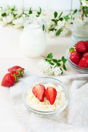 Bowl of cottage cheese, strawberry, cup of milk and white flowers on a white wooden background. Healthy breakfast. Diet. Vertical shotの写真素材