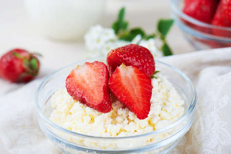 Close view at bowl of cottage cheese, strawberry, cup of milk and white flowers on a white wooden background. Healthy breakfast. Diet.の写真素材