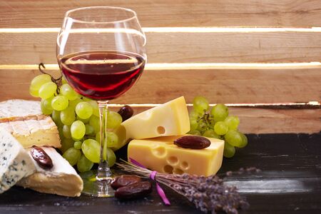The Glass of red wine served with grapes, date fruits and the variety of cheese on a black wooden table with the wooden board on a background with a ray of light shining through it. Close viewの写真素材