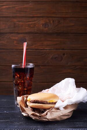 Small graceless burger on a paper package served with a glass full of ice and drink. At a dark wooden background. Close view. Vertical shotの写真素材