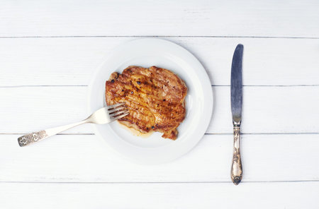 Delicious beef steak on white plate with fork and knife on white wooden background with space for...