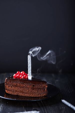 A slice of chocolate cake with redcurrant and a single extinguished candle on a dark background. Vertical shotの写真素材