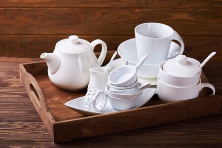 Set of new white dishes with teapot, tea cups, and plates on wooden tray at a wooden tableの写真素材