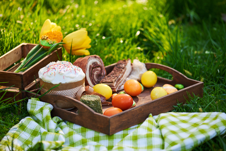 Easter cake and bright colored eggs gathered together with meat, treats and tulips on the lawn among the grassの写真素材