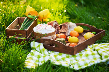 Easter cake and bright colored eggs gathered together with meat, treats and tulips on the lawn among...