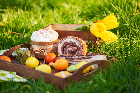 Easter cake and bright colored eggs gathered together with meat, treats and tulips on the lawn among the grassの写真素材